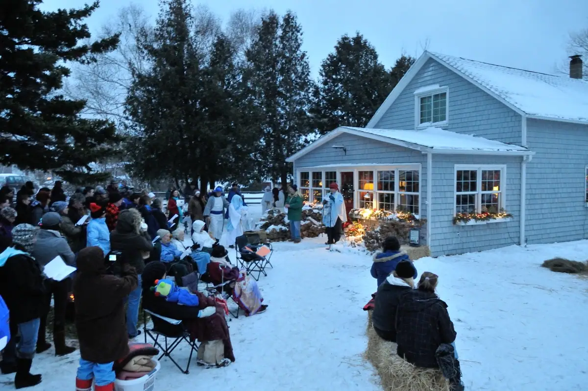 A winter crowd watches costumed participants gathered beside a snow-covered house decorated with lights.