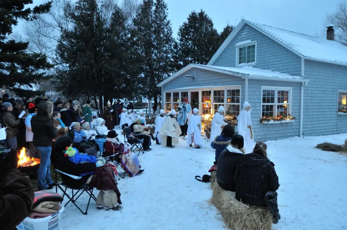 People in white robes perform outdoors in the snow while an audience watches beside a blue-shingled house.