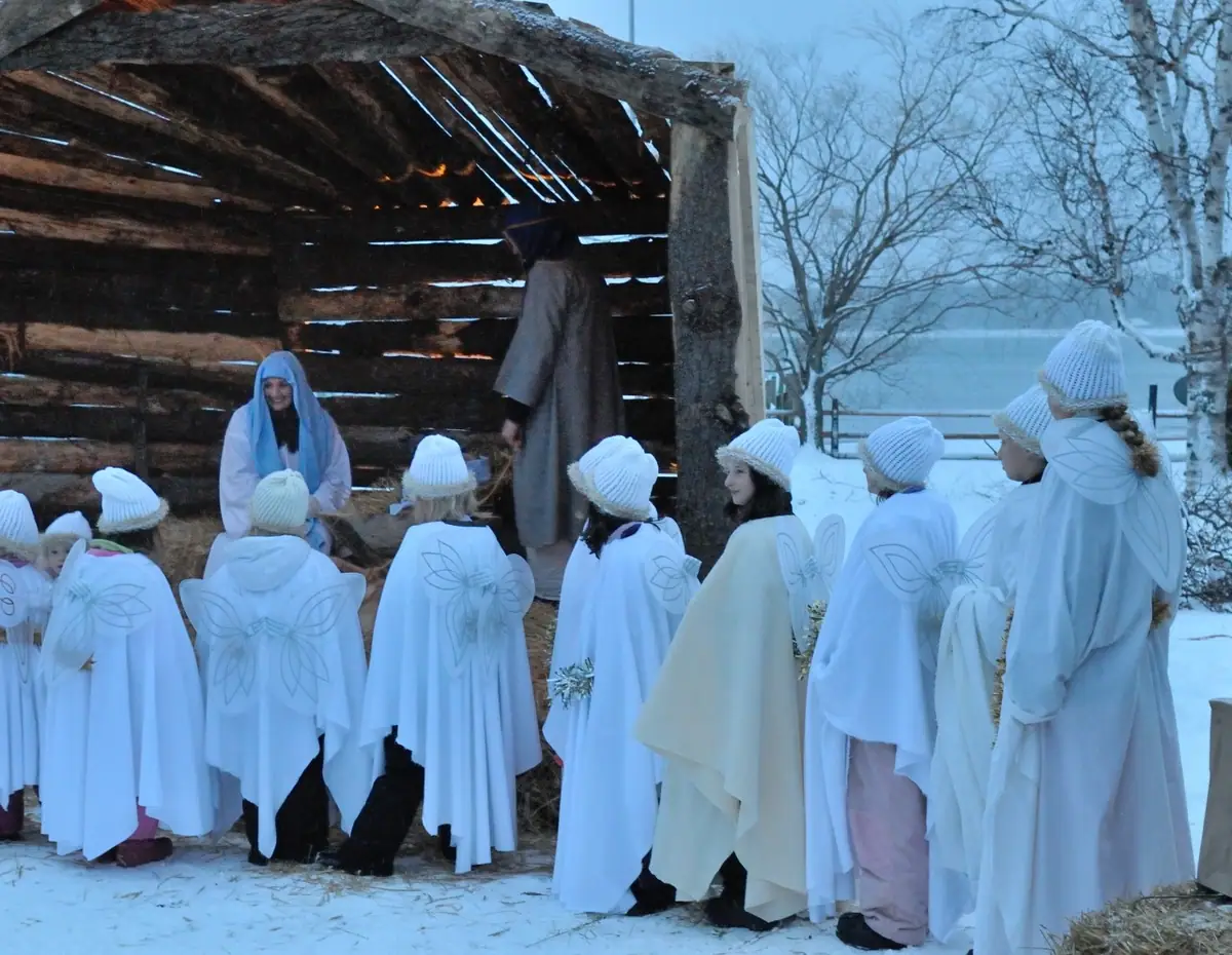 Children in white angel costumes gather around a rustic wooden shelter in a snowy outdoor nativity scene.