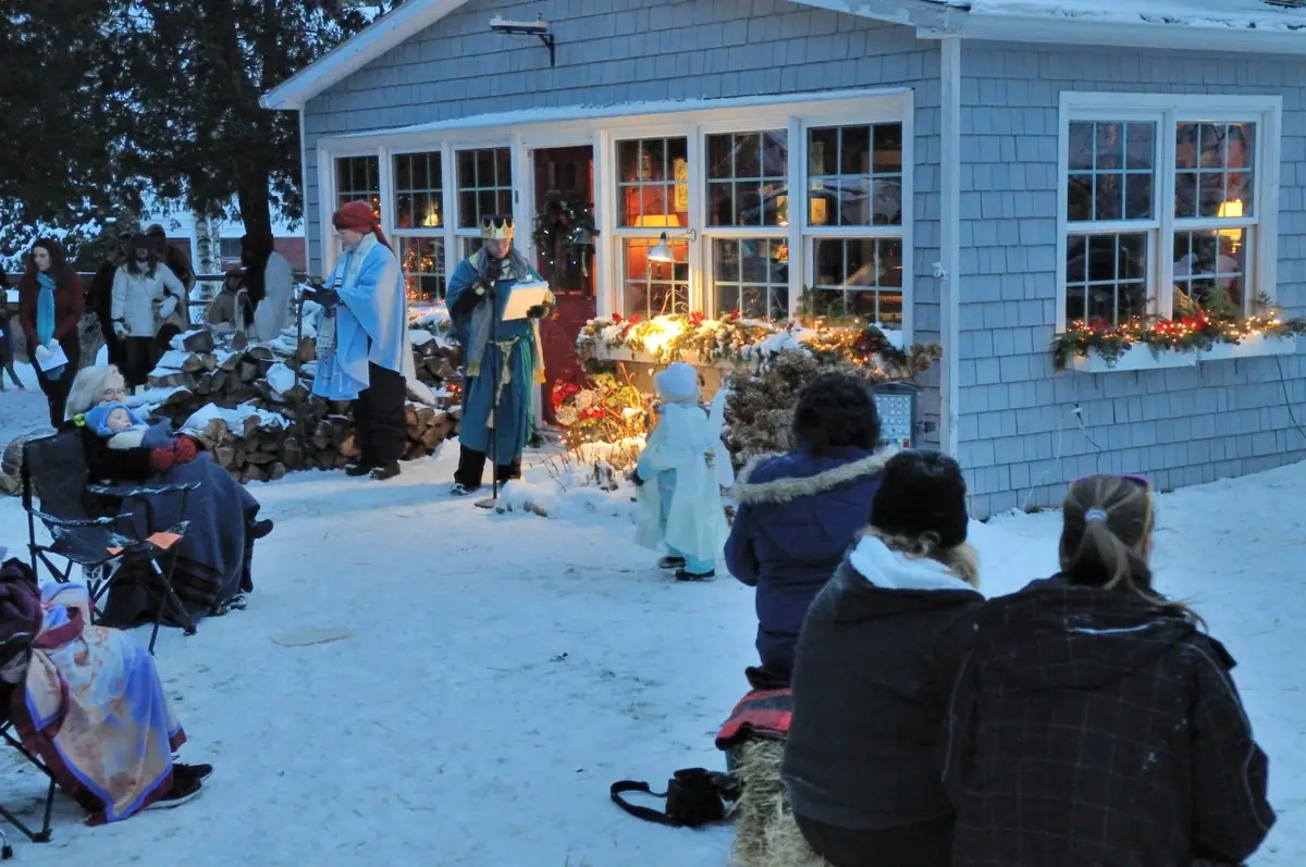 A winter nativity performance unfolds outside a shingled house as spectators watch.