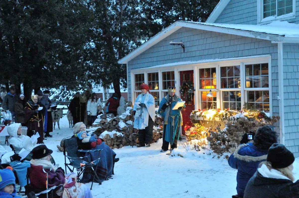 A costumed outdoor nativity scene unfolds in the snow beside a blue-shingled building as spectators watch.