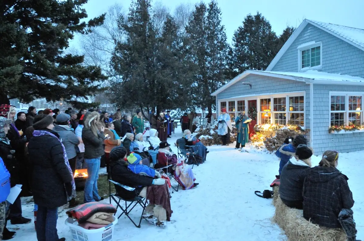 A winter crowd watches costumed figures outside a shingled house, with a small fire glowing among the spectators.