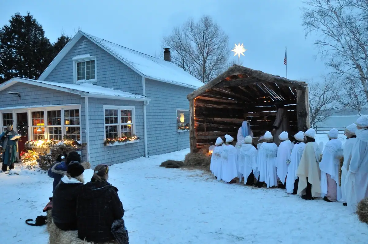 People in white robes gather beside a lit wooden nativity shelter in a snowy yard.
