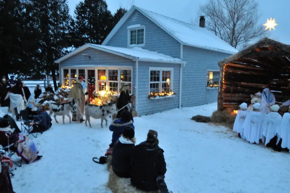 People in robes and winter clothing gather with animals beside a lit stable and blue-shingled house in the snow.
