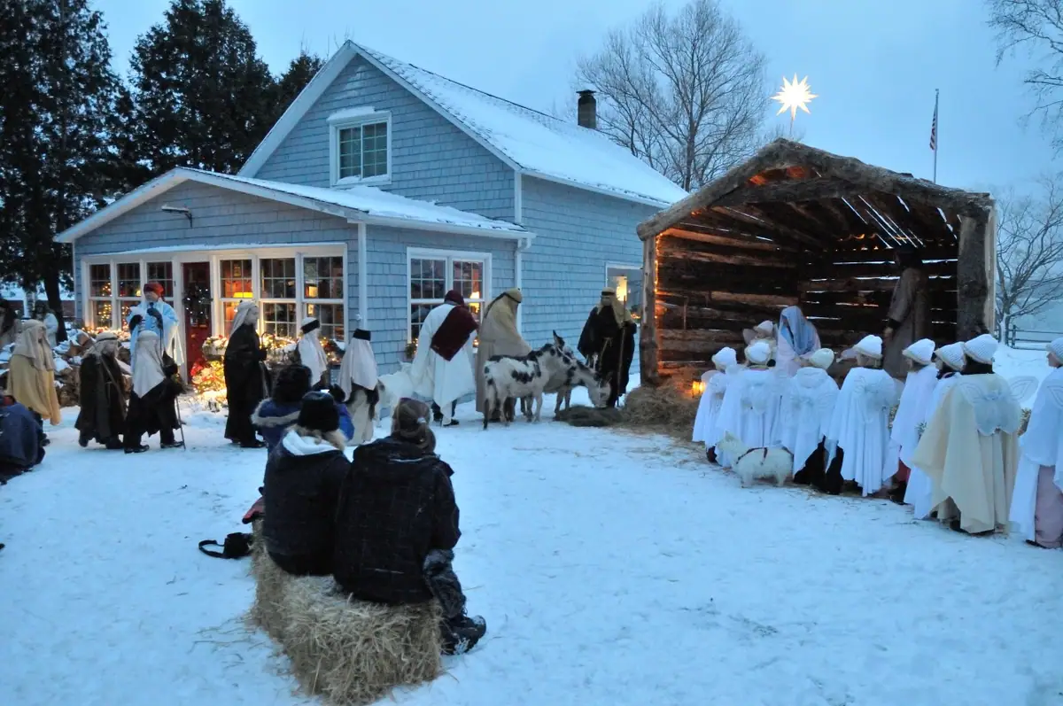 Outdoor nativity scene with costumed people, goats, sheep, a stable, and a star beside a snowy house.
