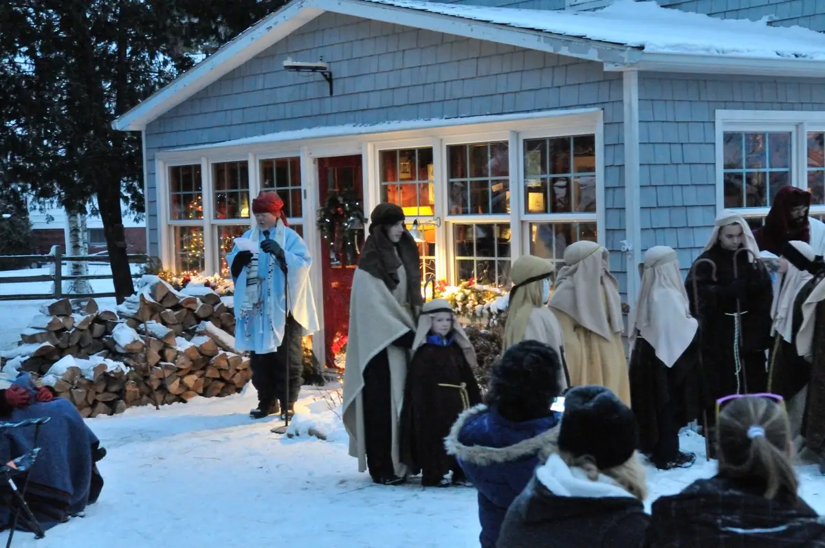 Costumed performers and an audience gather in the snow outside a shingled house.