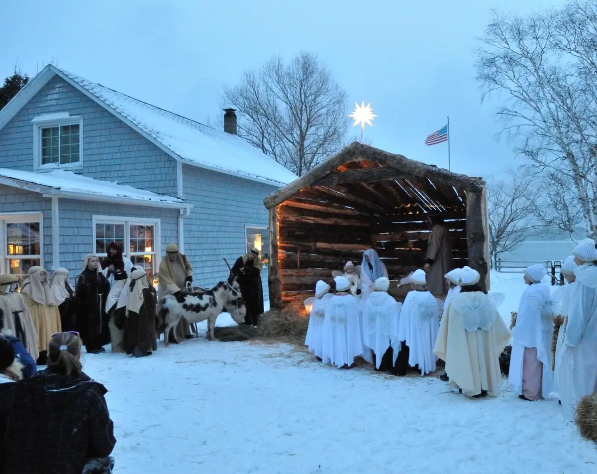 Costumed participants and a goat gather beside a lit wooden shelter in a snowy yard.