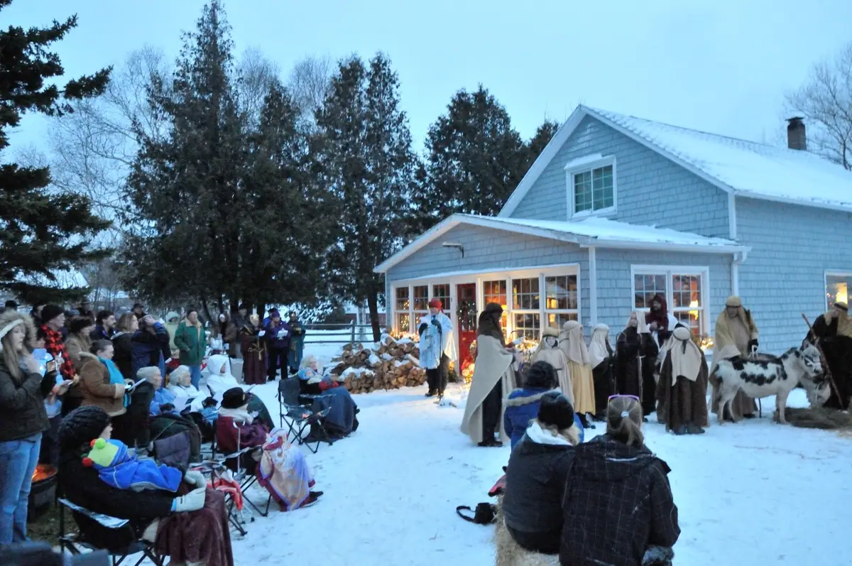 A costumed nativity scene unfolds beside a snow-covered house as spectators watch.