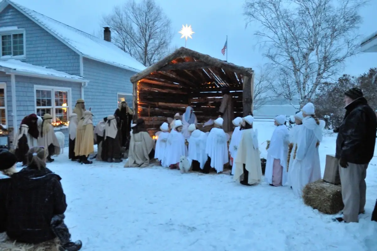 People in robes gather around a lit wooden shelter in a snowy yard during a staged nativity scene.