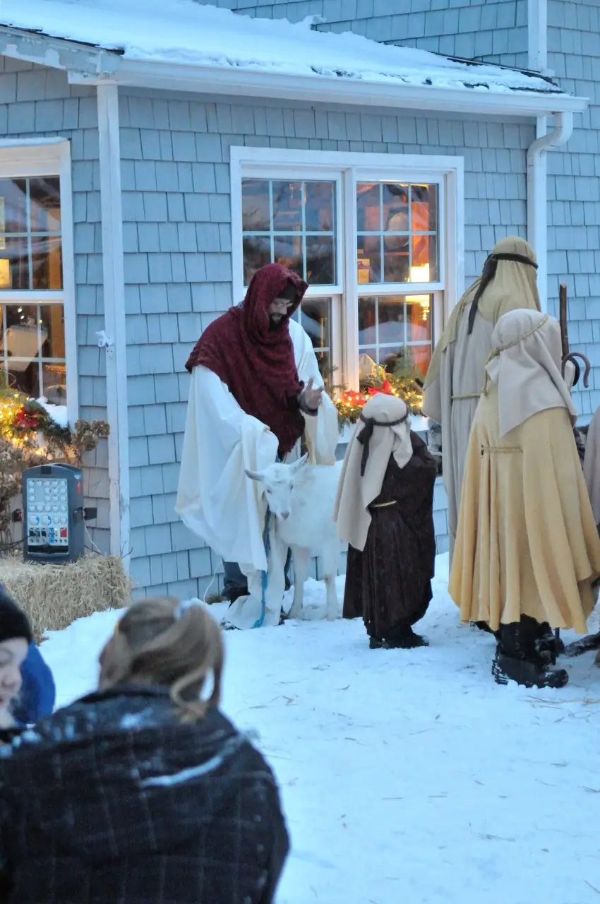 Costumed figures and a goat perform outdoors in snow as children watch beside a blue-shingled house.
