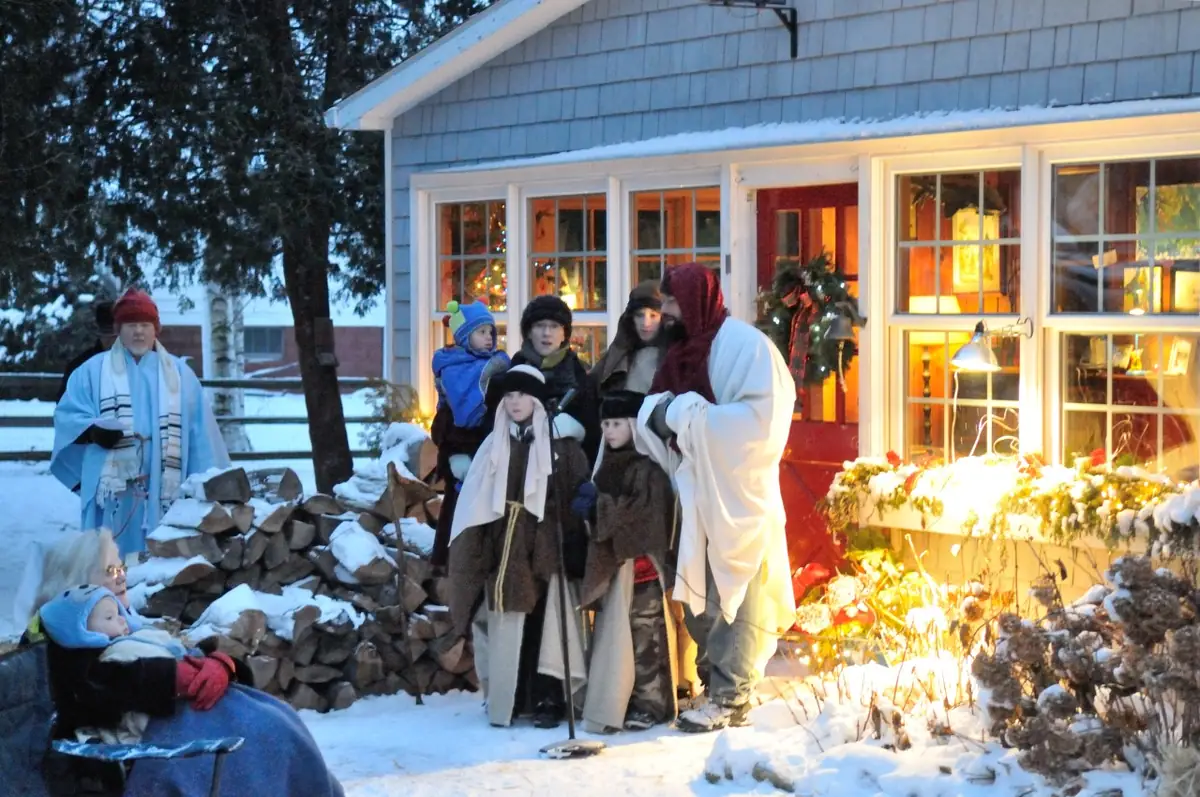 People in robes and winter clothing gather in snow outside a decorated house at twilight.