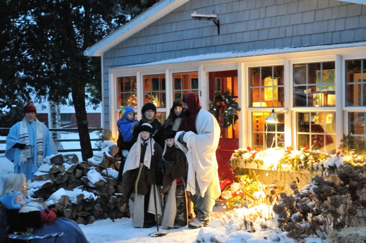 People in winter clothing and robes gather outside a brightly decorated, snow-covered house.