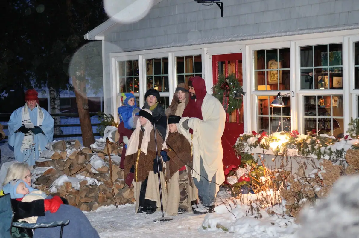 People in robes and winter clothing gather for an outdoor performance in snowy surroundings beside a decorated house.