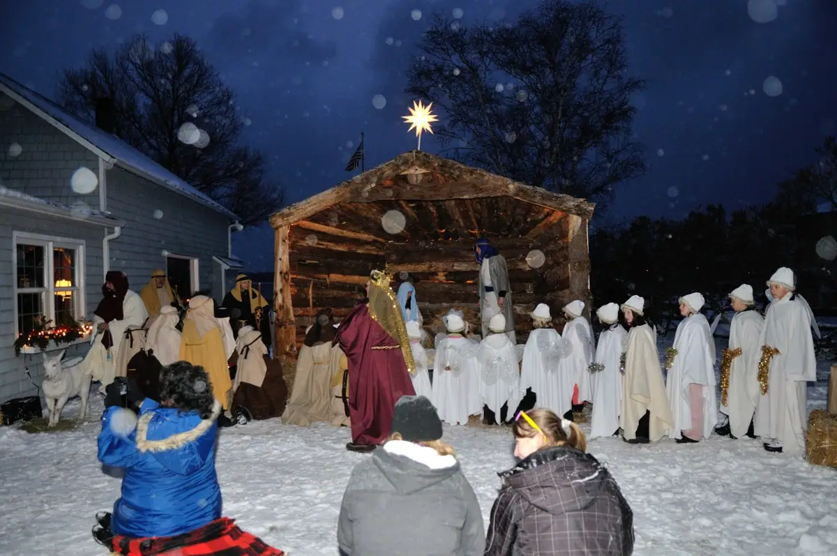 People in winter costumes gather around a rustic stable during an outdoor nativity performance at night.