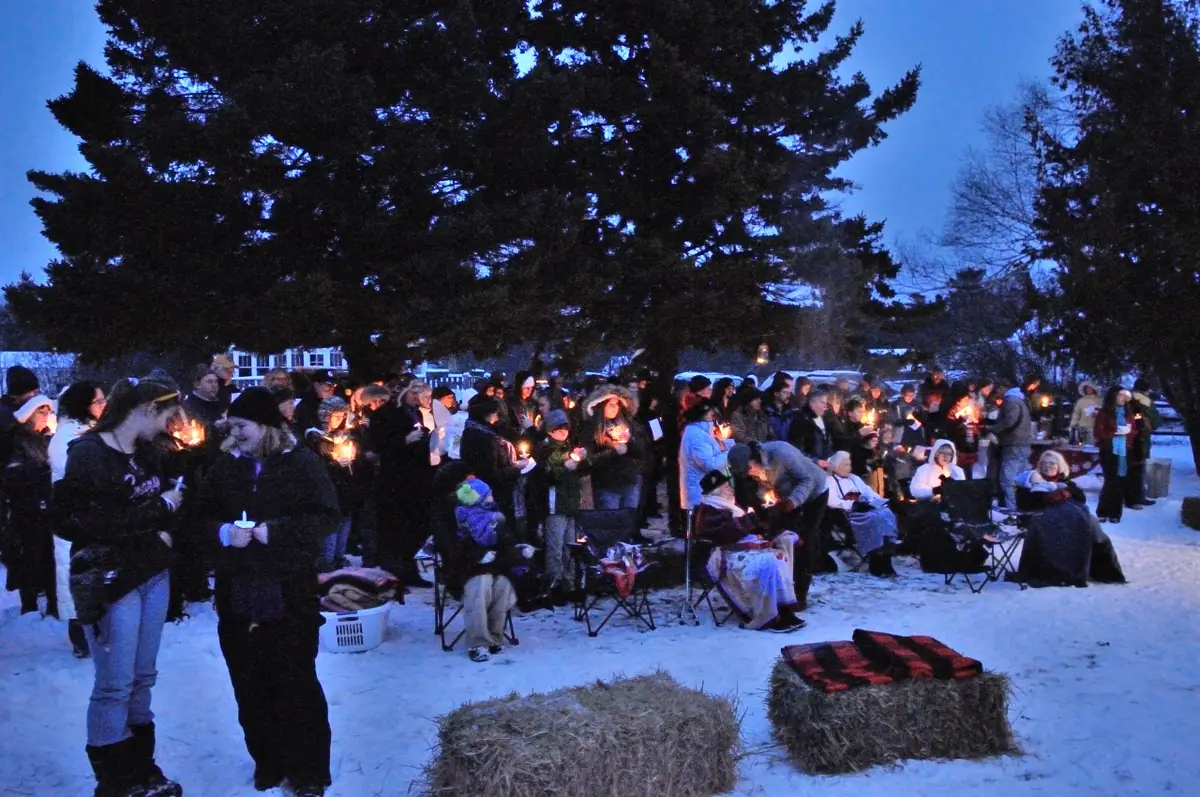 A candlelit crowd gathers in a snowy outdoor setting beside hay bales and evergreen trees.