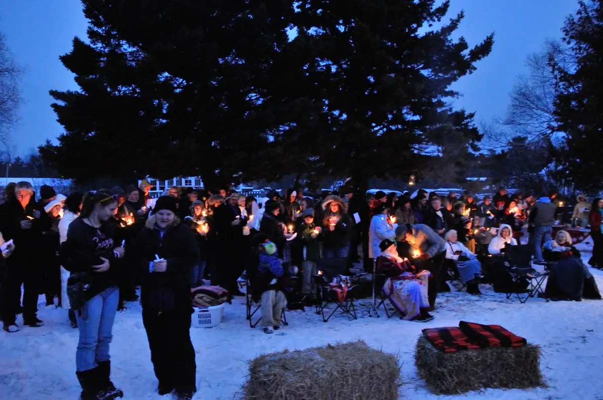 A candlelit crowd gathers in the snow beside hay bales at dusk.