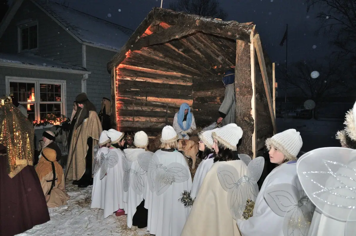 Children in angel costumes gather before a lit wooden shelter during a snowy nighttime nativity scene.