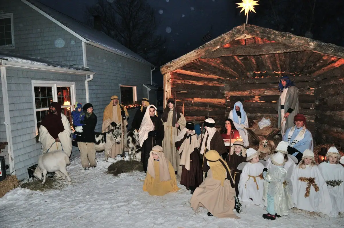 People and children in robes gather with goats beside a rustic stable in a snowy yard at night.