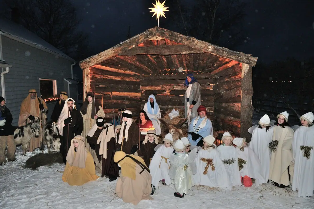 Costumed adults and children gather around a lit log shelter in a snowy nighttime nativity scene.