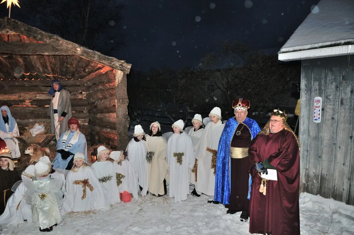 People in nativity costumes and white robes gather in the snow beside a rustic stable at night.