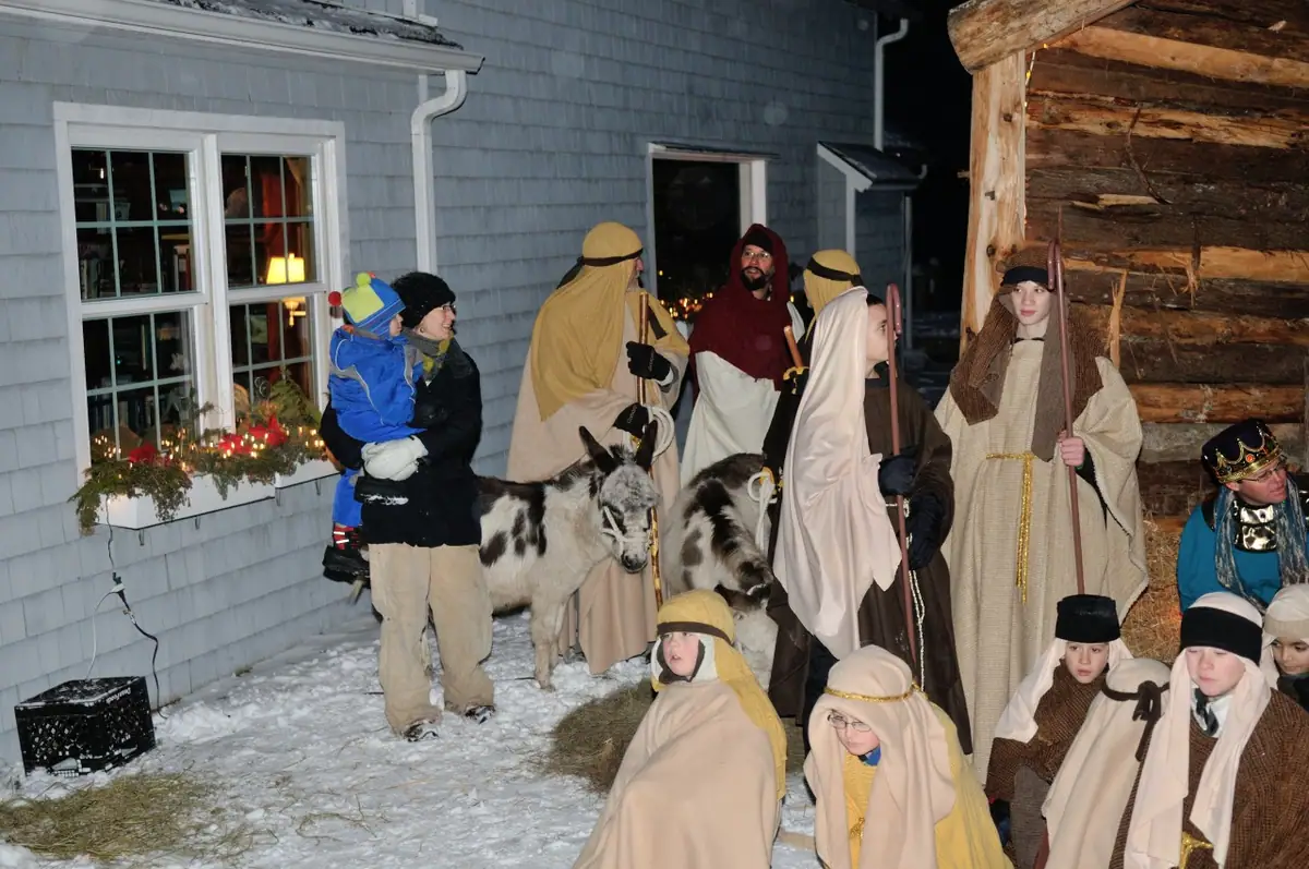 Costumed adults and children gather with two donkeys beside a snowy, illuminated rustic shelter.