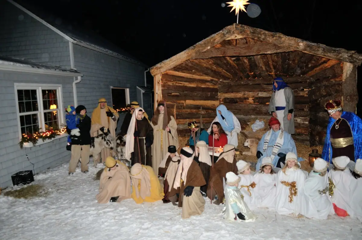 A costumed group gathers around a rustic wooden shelter in a snowy outdoor nativity scene at night.