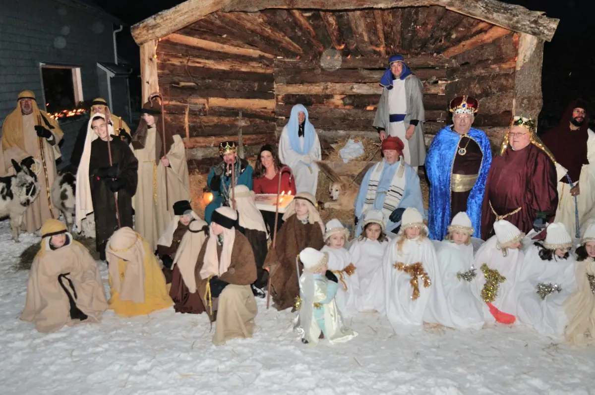 Adults and children in nativity costumes pose before a rustic log shelter in the snow at night.
