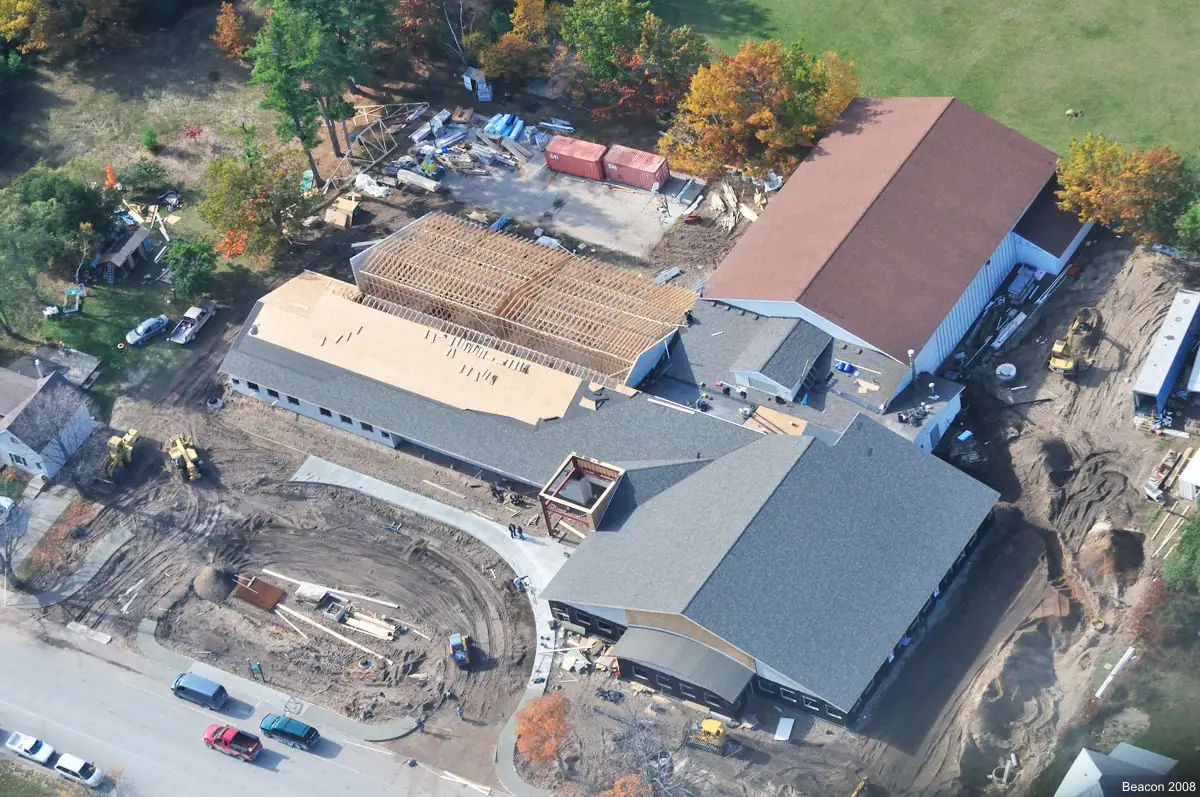 Aerial view of a large building complex under construction, with exposed roof framing, construction equipment, and autumn trees.