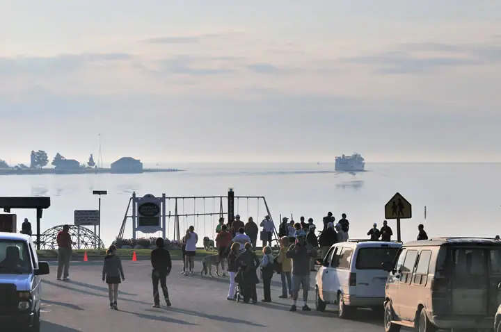People gather beside a playground and waterfront as a ferry sits offshore.