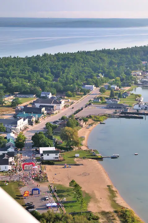Aerial view of a shoreline town, beach, docks, and a large crowd gathered beside colorful event structures.