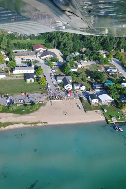 Aerial view of a beach, race gathering, streets, buildings, and turquoise water beneath an airplane wing.