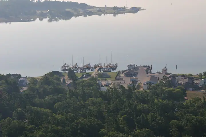 Aerial view of a marina, sailboats, waterfront buildings, wooded shoreline, and a distant tower.
