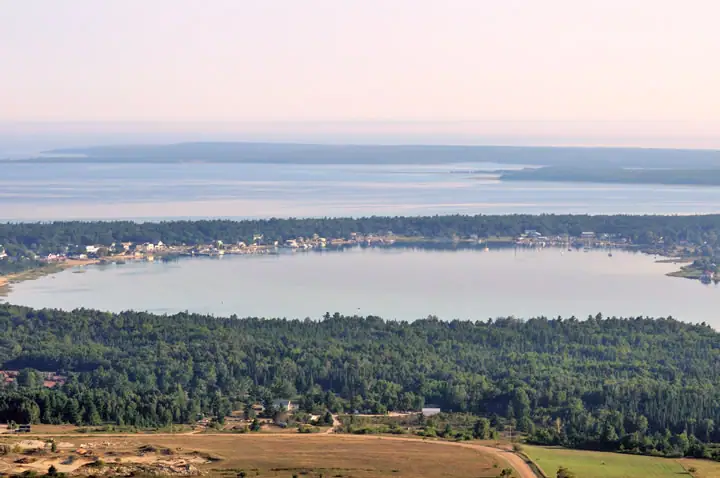 Aerial view of a wooded shoreline, a broad calm inlet, waterfront homes, fields, and a winding road.