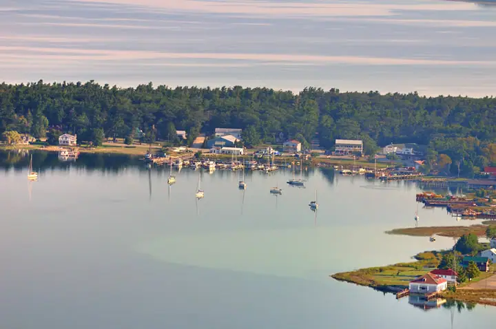Aerial view of a calm harbor with moored sailboats, docks, houses, and a wooded shoreline.