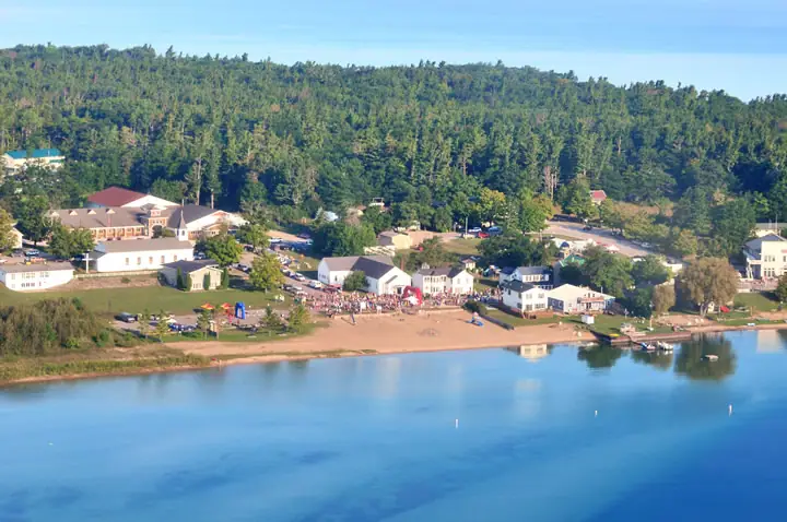 Aerial view of a shoreline settlement with a sandy beach, gathered people, buildings, and a wooded hillside.