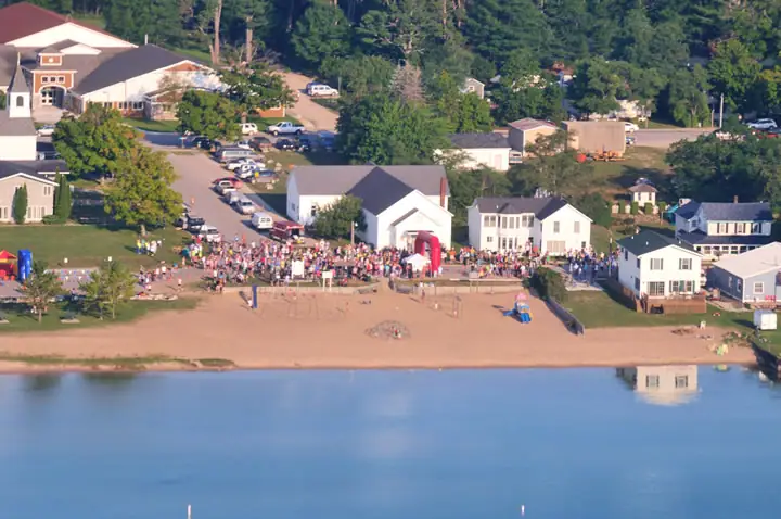 Aerial view of a crowd gathered beside a sandy shoreline, with a red inflatable arch and nearby homes.