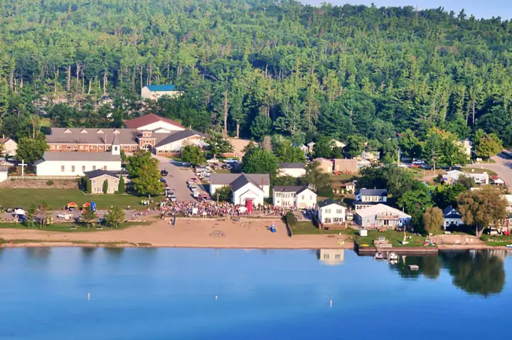 Aerial view of a waterfront settlement with a crowd gathered beside a sandy shore and wooded hillside.