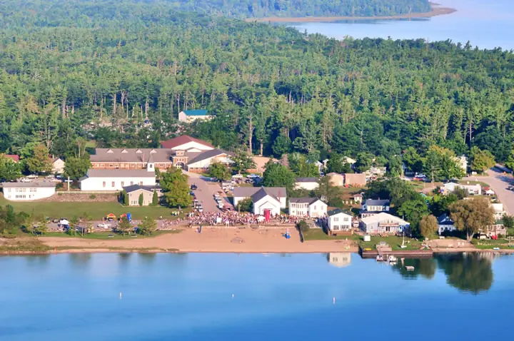 Aerial view of a waterfront settlement with a crowd gathered along the beach and forested land beyond.