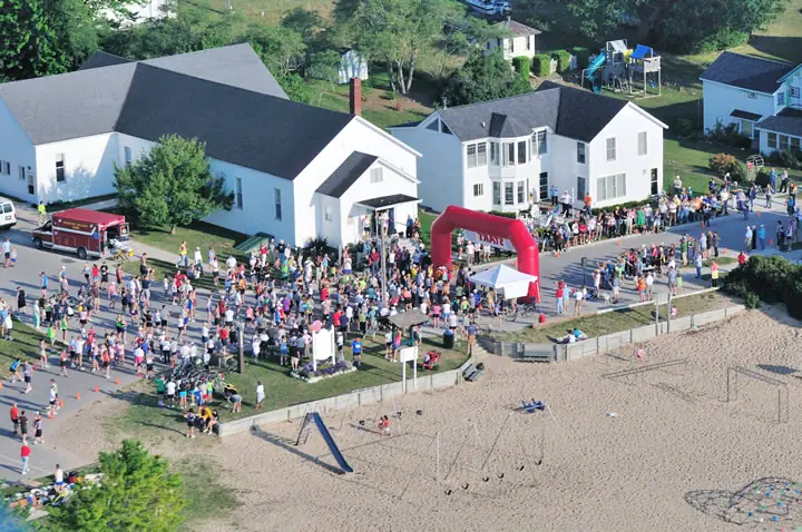 Aerial view of a crowded road race gathering beside a sandy beach, with an inflatable arch and nearby homes.