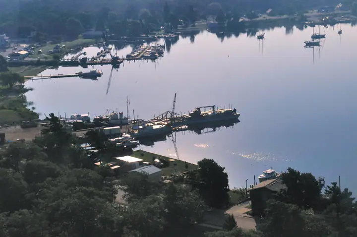 Aerial view of a harbor with workboats, docks, cranes, sailboats, shoreline buildings, and wooded surroundings.