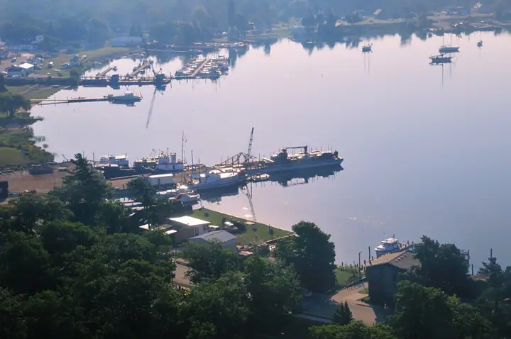 Aerial view of a harbor with docks, boats, a work vessel, buildings, and wooded shoreline.