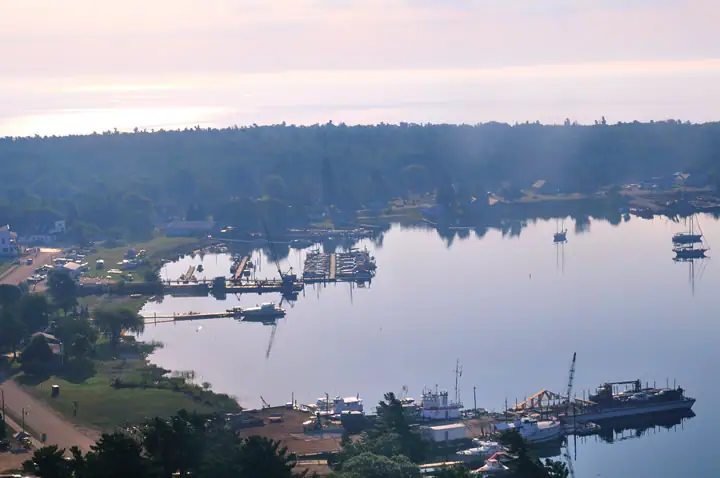 Elevated view of a harbor with docks, boats, a wooded shoreline, and a road.