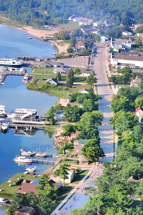 Aerial view of a waterfront town with a harbor, ferry, boats, streets, trees, and people along a roadway.