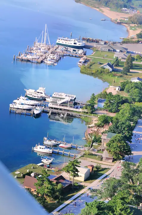 Aerial view of a marina with a ferry, sailboats, docks, waterfront buildings, and people along a road.