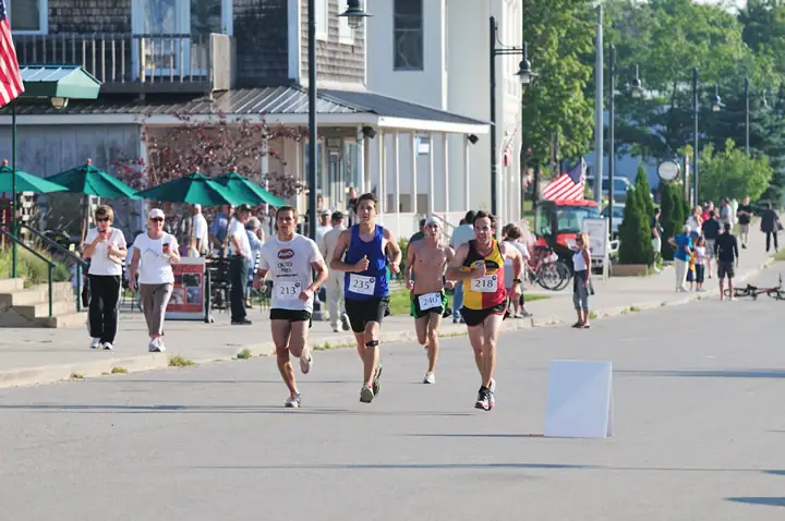 Four runners compete on a paved street lined with storefronts, spectators, umbrellas, and flags.