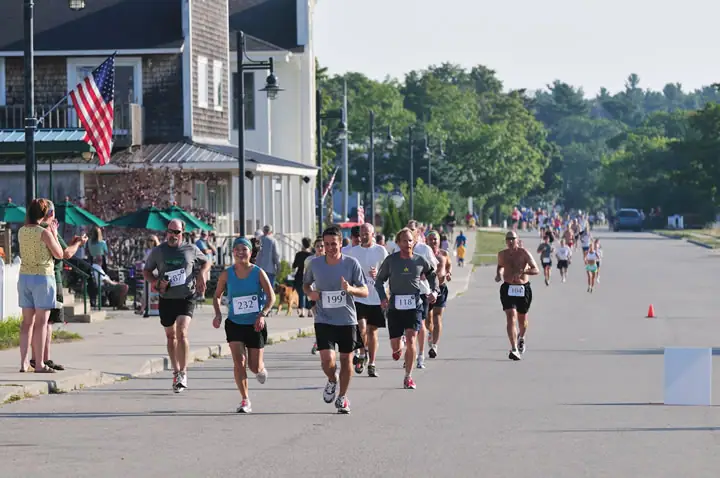 Runners in numbered bibs compete along a broad street as spectators watch from the sidewalk.
