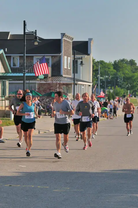 Runners move along a paved street past storefronts, spectators, hanging lamps, and an American flag.