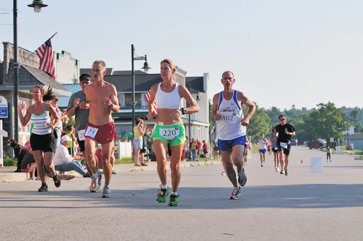 Runners wearing numbered bibs compete along a paved street, with an American flag and spectators behind them.