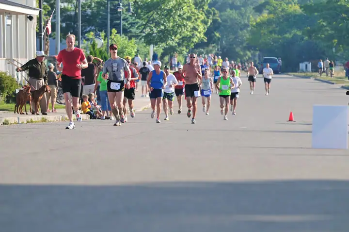 Runners advance along a paved street while spectators and a dog watch from the roadside.