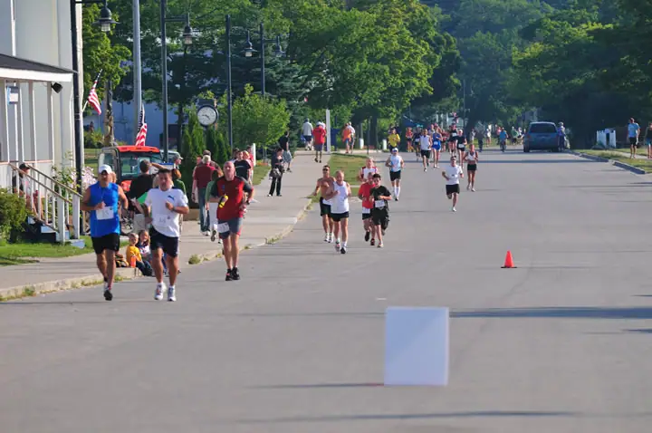 Runners move toward the camera along a broad paved street lined with trees, buildings, flags, and a traffic cone.